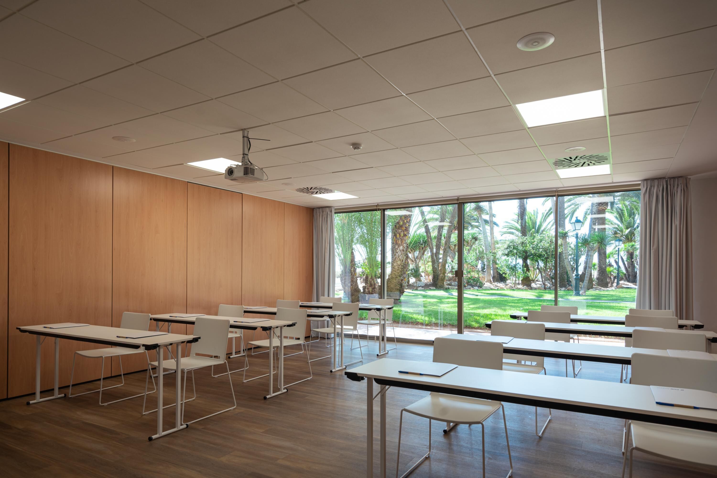 Un'aula con file di banchi e sedie, ampie finestre e vista su un giardino.