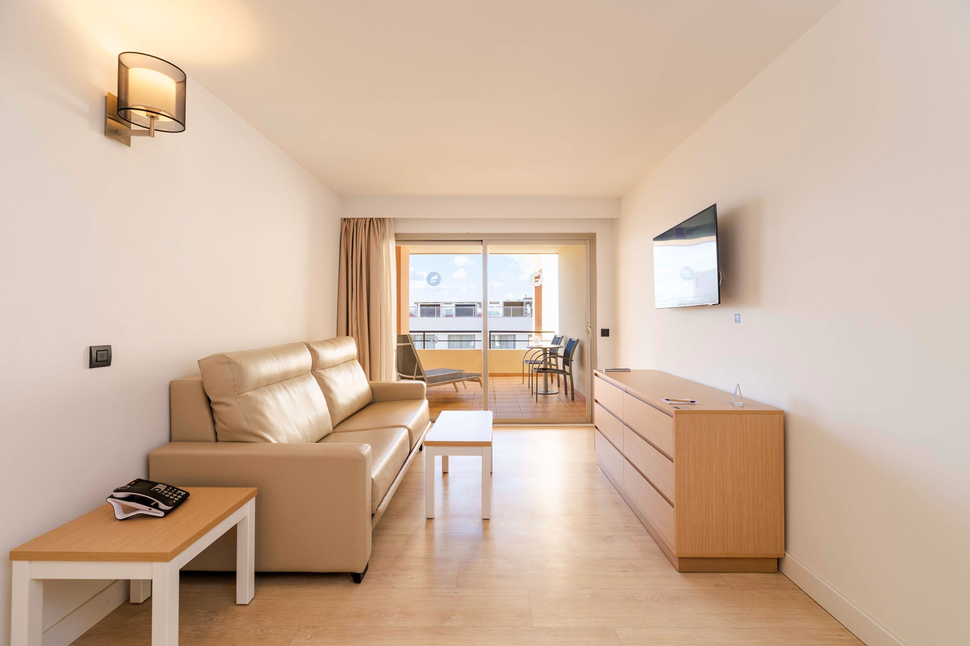 A modern living room with a sofa, a small table, a TV mounted on the wall, and a balcony with a view.