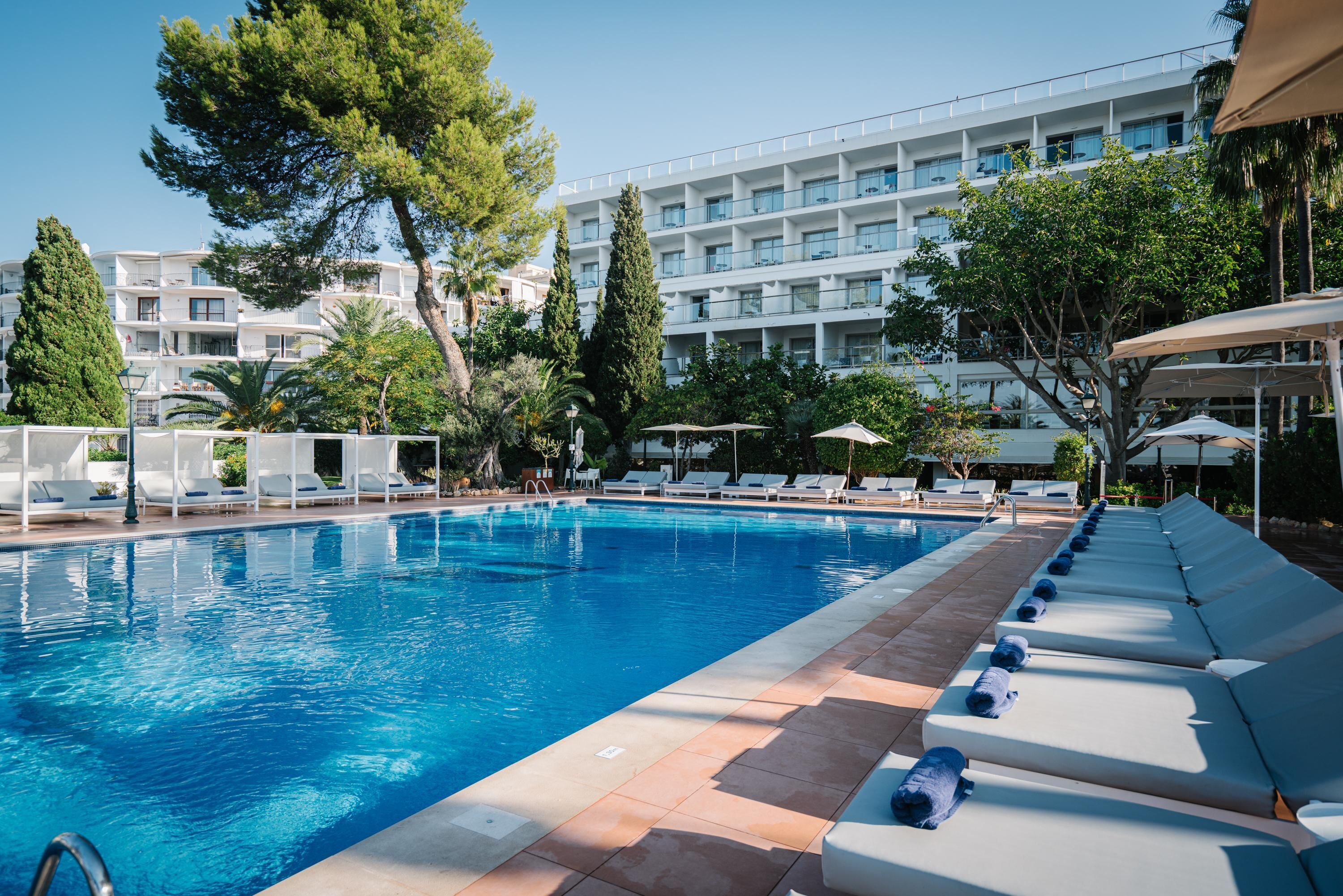 A swimming pool with lounge chairs and umbrellas, surrounded by trees and a multi-story building.