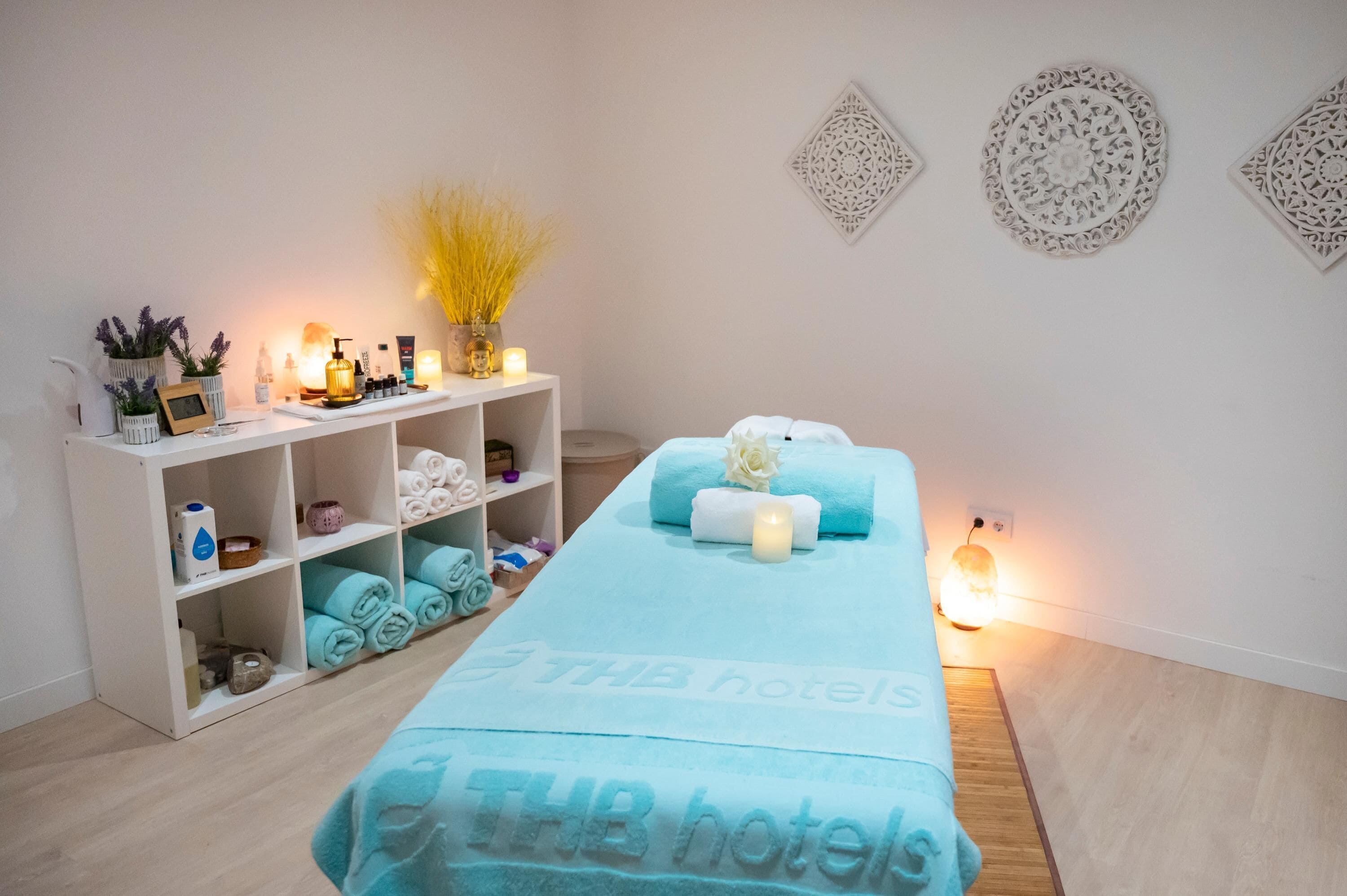 A spa room with a massage table, shelves with towels and toiletries, decorative wall art, and a small plant.
