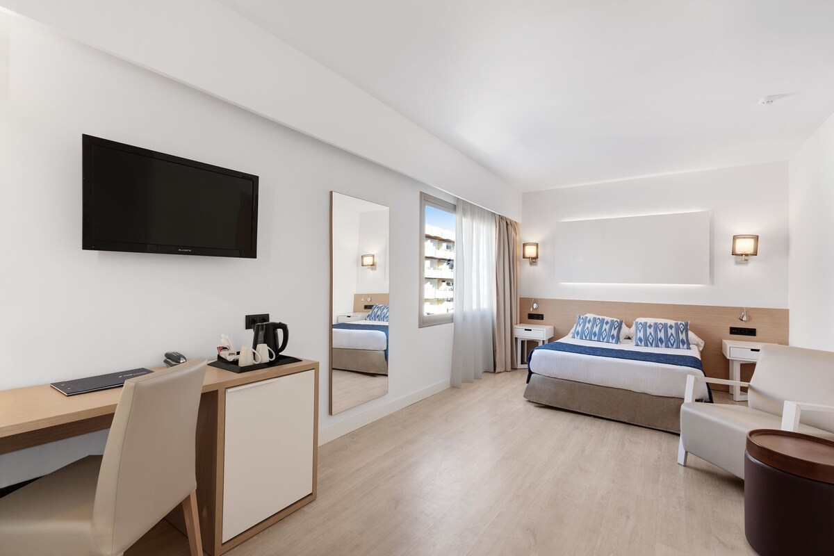 Una habitación de hotel moderna con televisión de pantalla plana, un escritorio con computadora, una cama con cabecera, una mesita de noche y una silla.