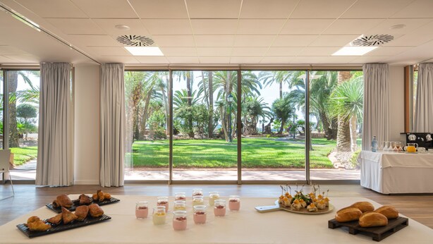 Amplia habitación con ventanas grandes que ofrecen vistas a un jardín bien cuidado con palmeras.