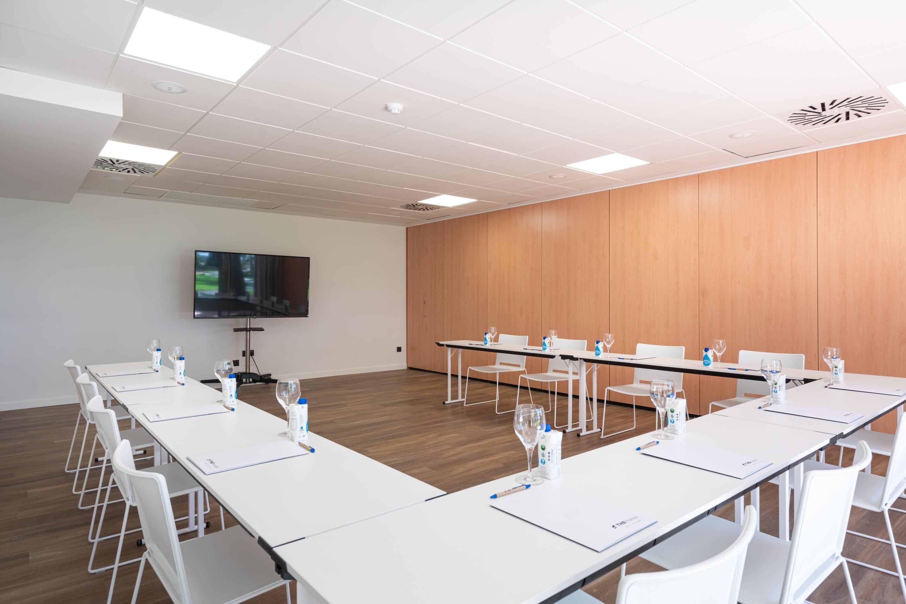 A conference room with white tables, chairs, and a flat-screen TV on the wall.