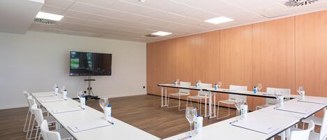 A conference room with white tables, chairs, and a flat-screen TV on the wall.