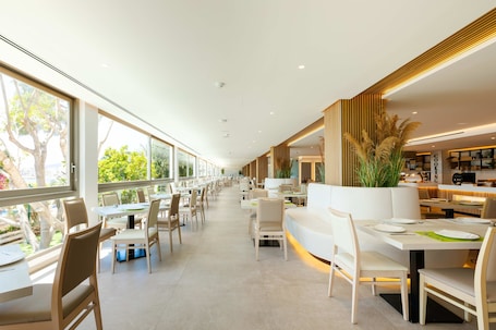 A modern restaurant with long tables and chairs, large windows, and a clean, minimalist interior.