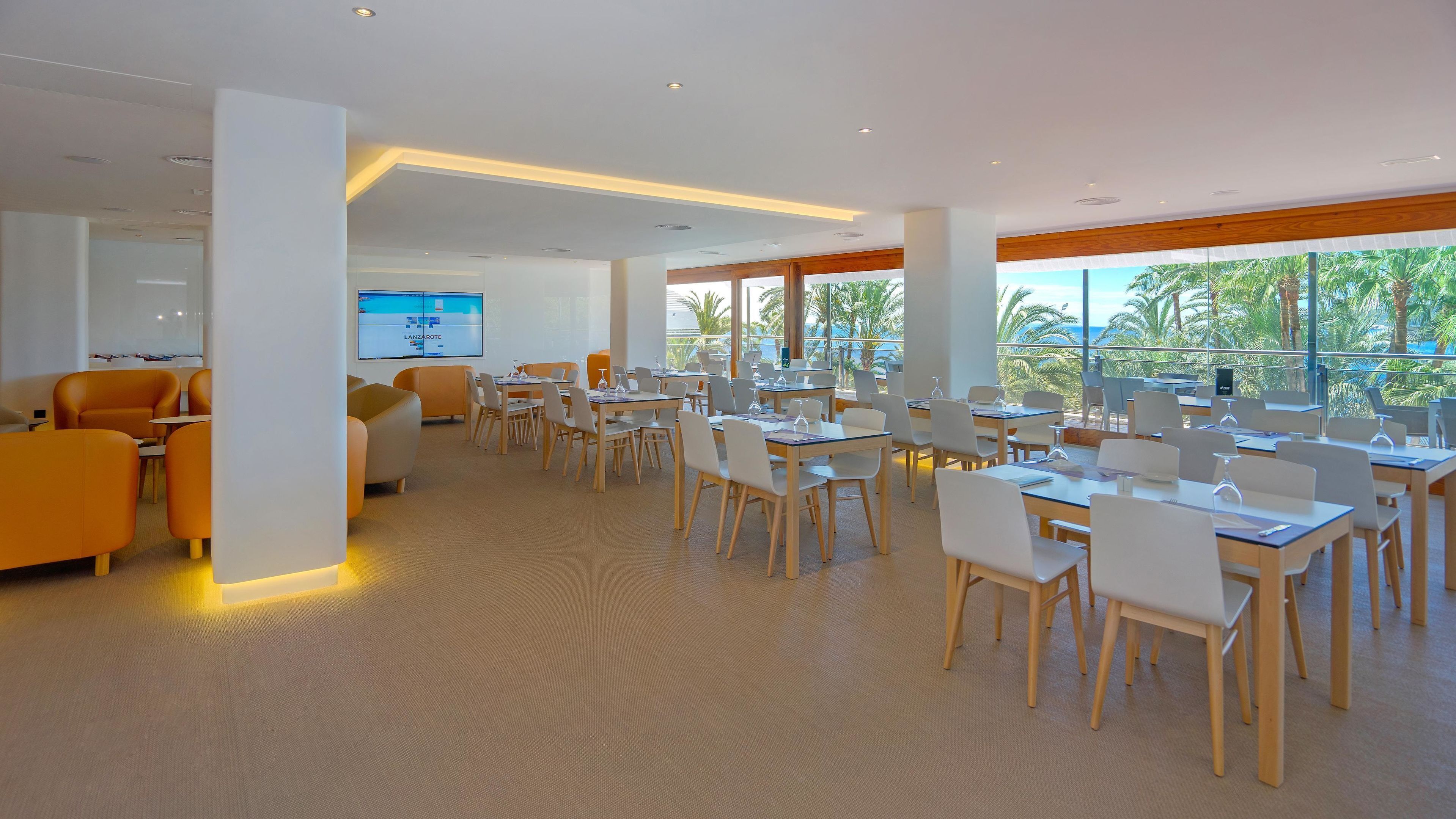 A modern conference room with white chairs and tables, large windows with a view of palm trees and the ocean, and a screen displaying a presentation.