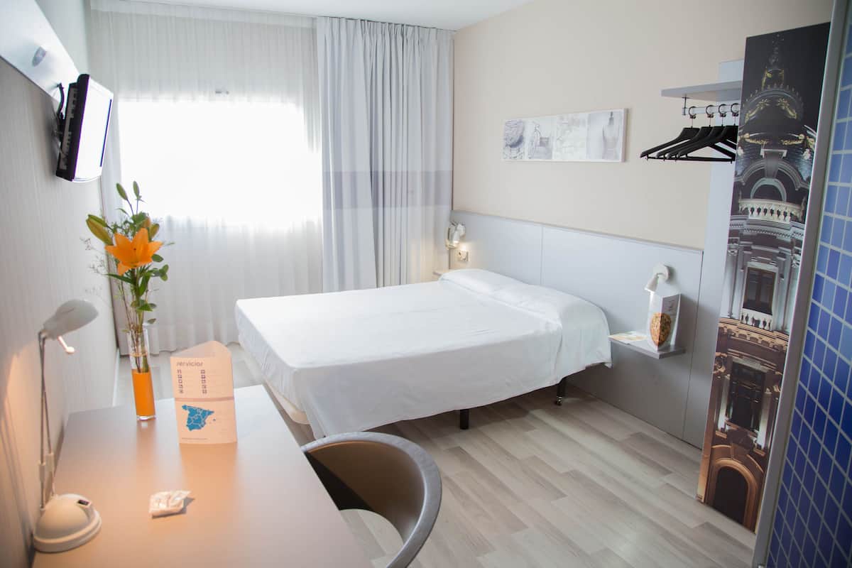 A hotel room with a bed, a desk with a lamp, a vase with flowers, a map, and a wall-mounted phone.