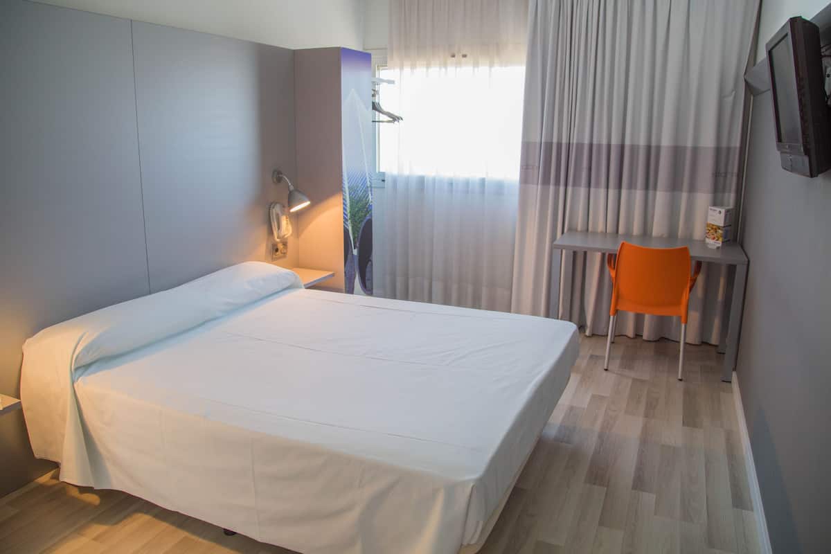 A hotel room with a bed, a desk, an orange chair, a TV, and a window with curtains.