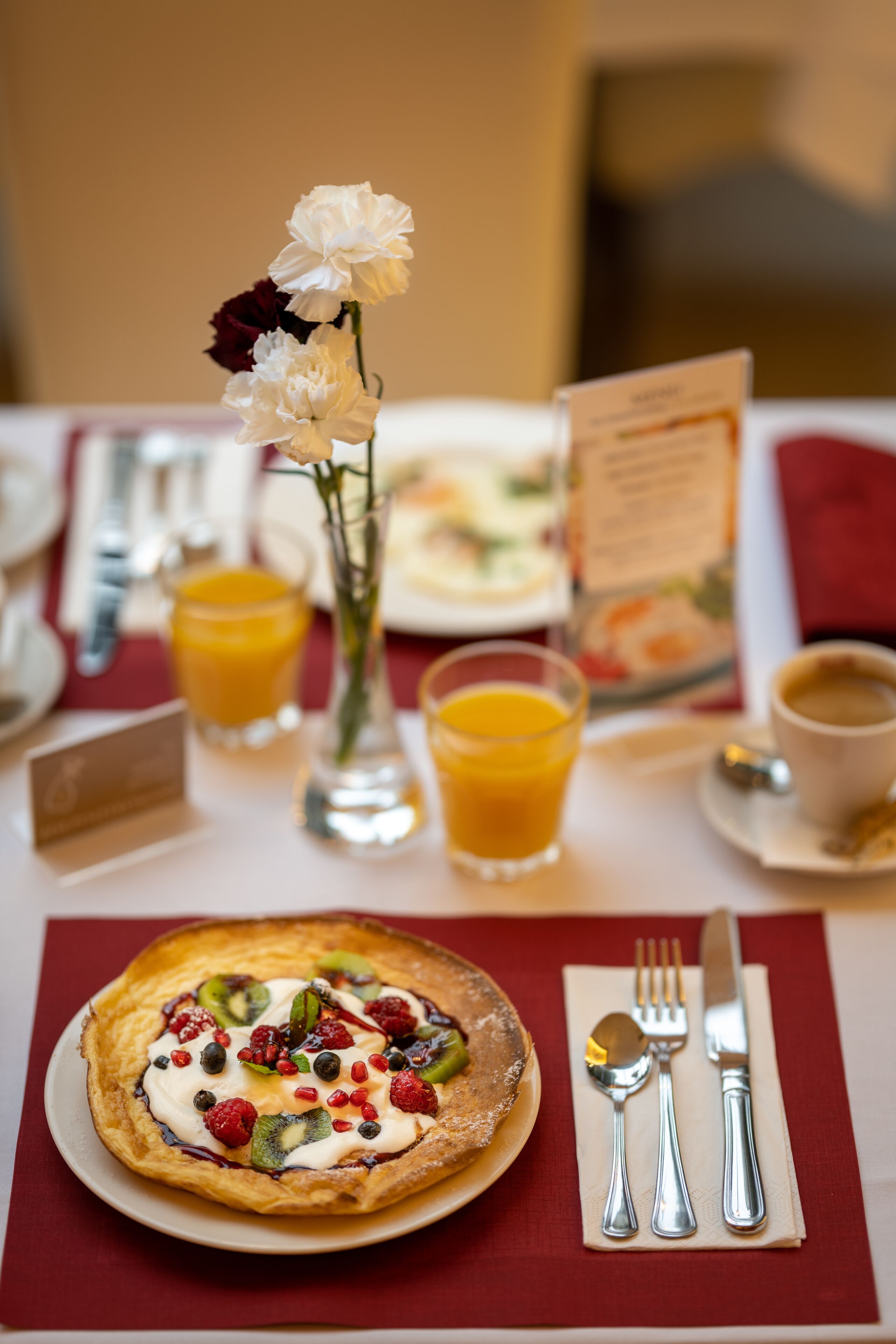 A breakfast setting with a plate of fruit-topped pastry, a glass of orange juice, a vase with flowers, and a menu.