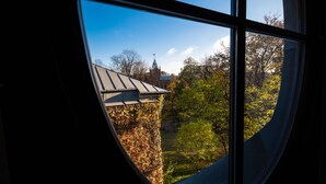 Uitzicht door een raam met een gebogen kader, waarop een gebouw met een puntige dak en een kruis te zien is, omgeven door bomen en groen.