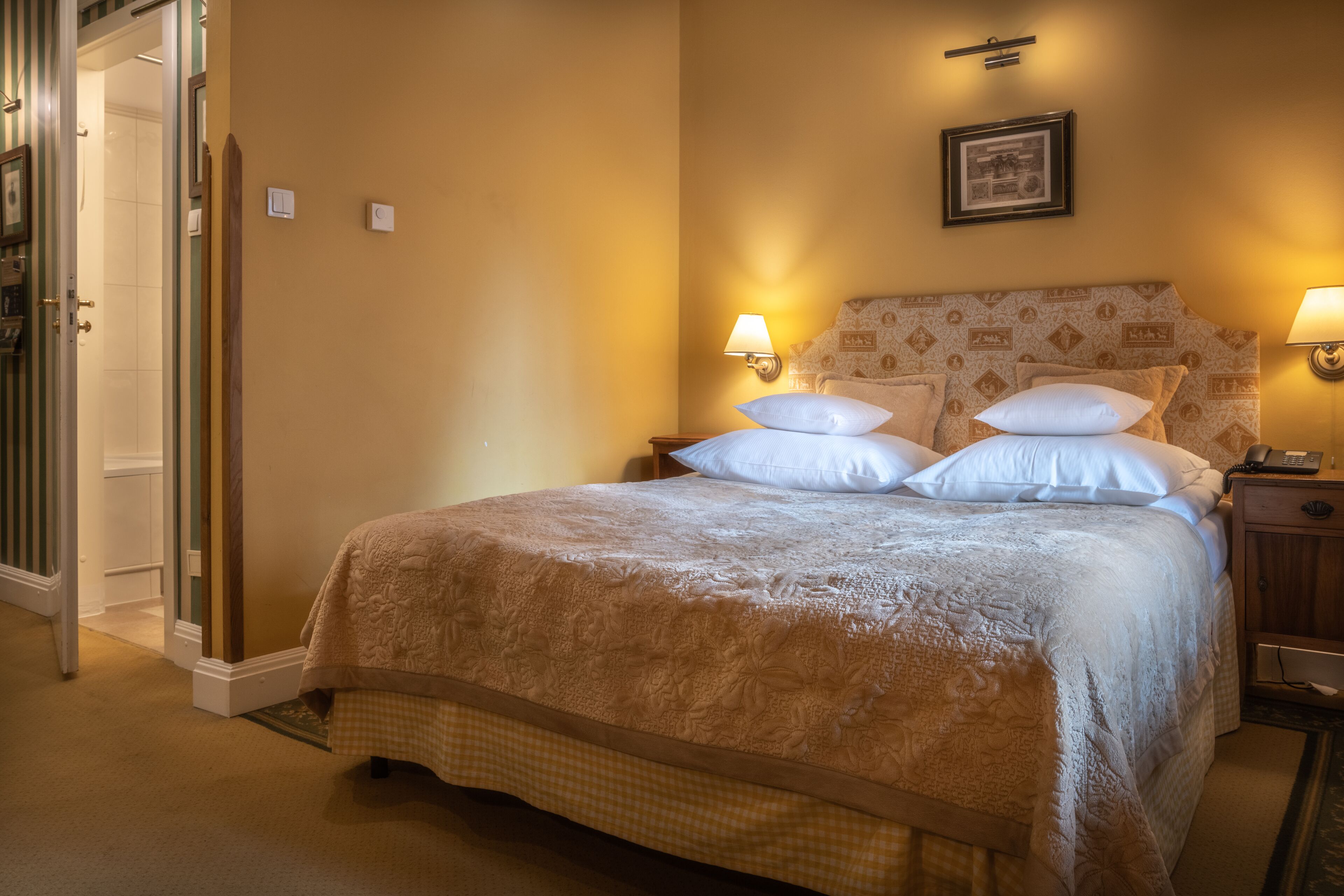 A hotel room with a large bed, two bedside lamps, a framed picture on the wall, and a door leading to another room.