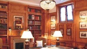 Een bibliotheek met houten boekenkasten, twee lampen, een open haard en ingelijste foto's aan de muren.