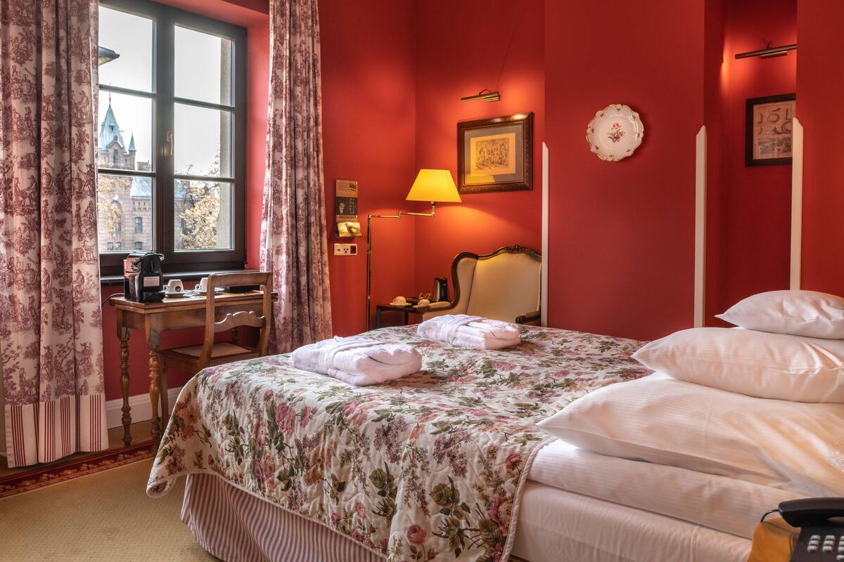 A hotel room with a large bed, a desk, a lamp, and a view of a building through a window.