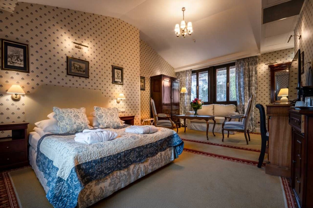 A bedroom with a large bed, a desk, and a chair. There are framed pictures on the wall and a chandelier above the bed.