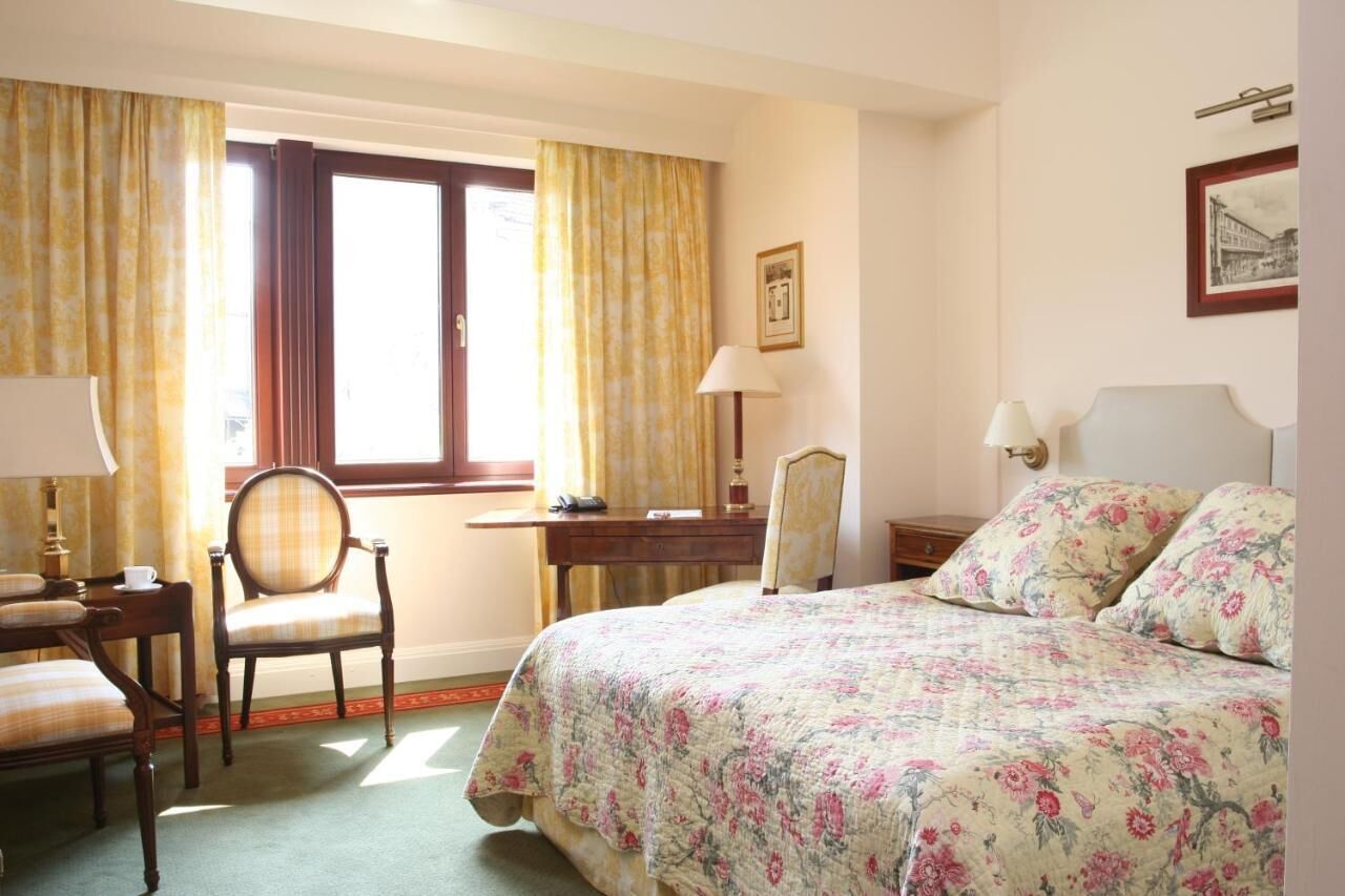 A hotel room with a bed, two chairs, a desk, a lamp, and a window with curtains.