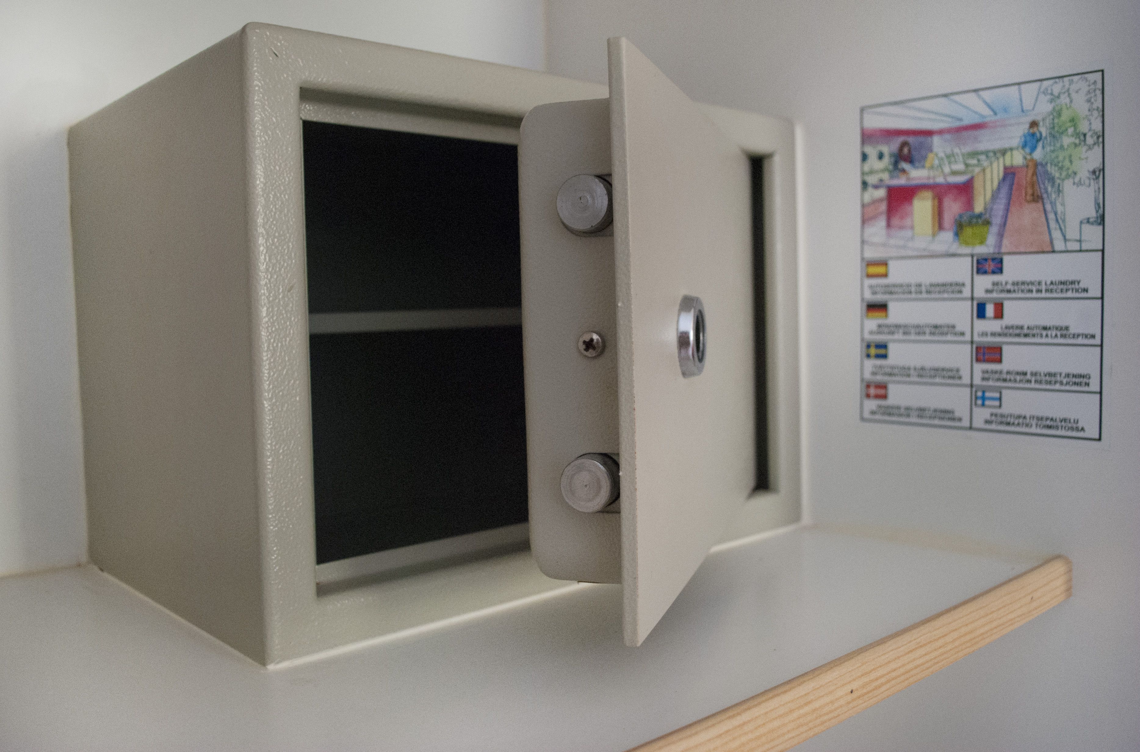 A beige safe with a keyhole and a metal latch, mounted on a wall with a sign displaying multilingual information.