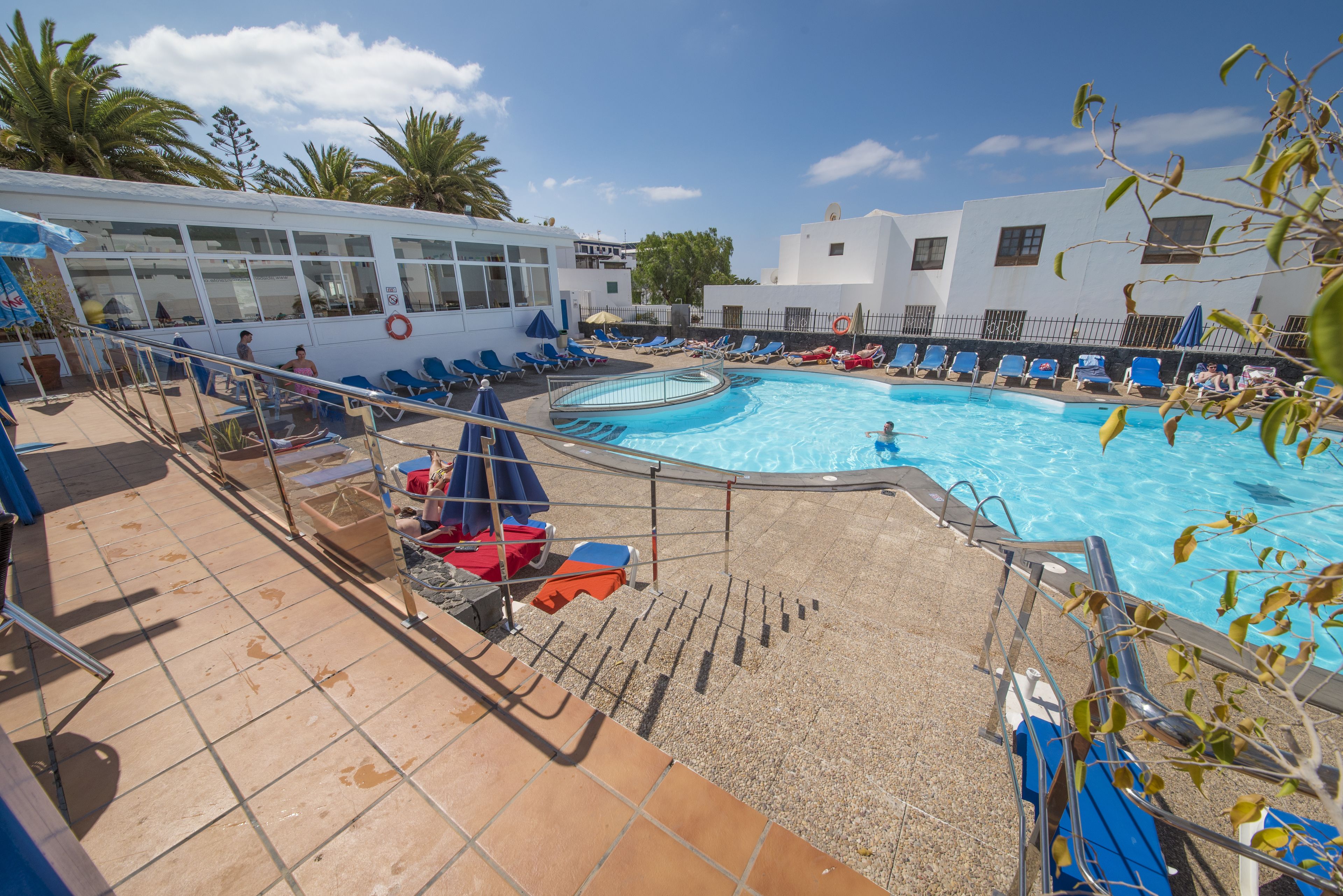 Un espace piscine doté de chaises longues et d’une bouée de sauvetage, entouré de palmiers et bordé d’un bâtiment aux grandes fenêtres.
