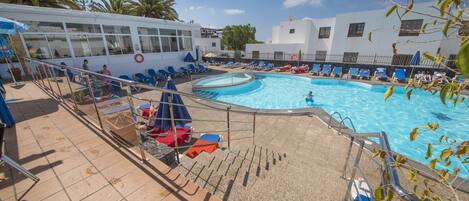 A pool area with sun loungers and a lifebuoy, surrounded by palm trees and a building with large windows.