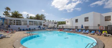 A swimming pool with lounge chairs and umbrellas, surrounded by palm trees and buildings.