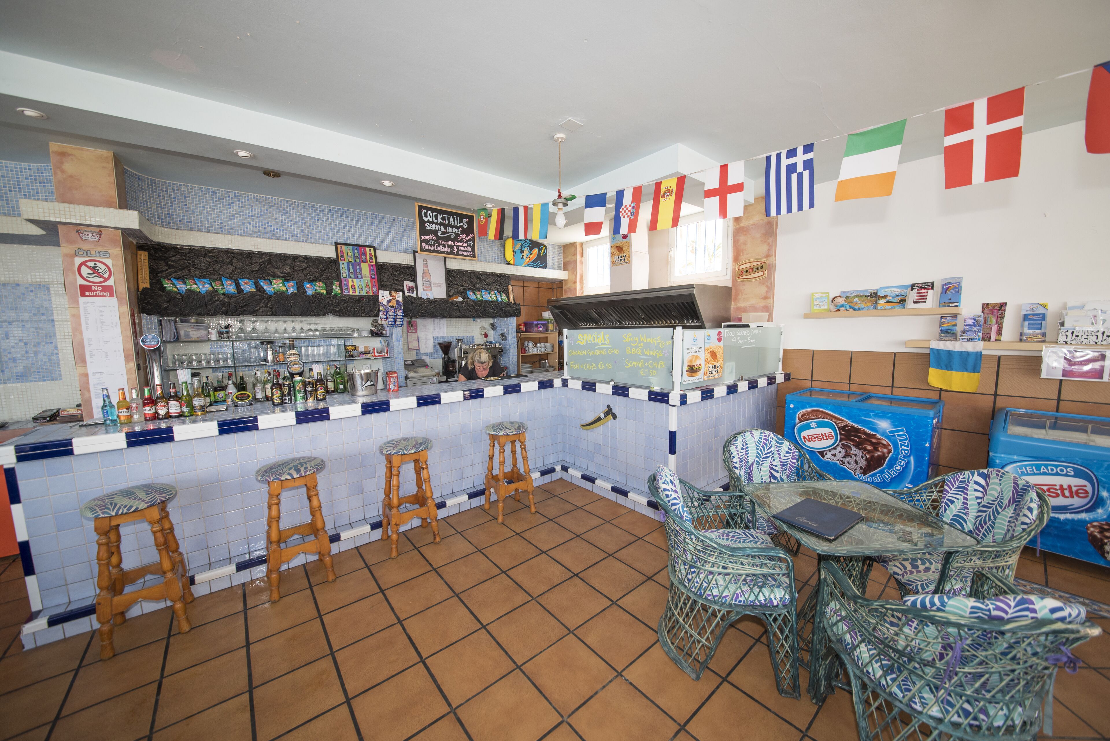 Een bar met krukken, een toonbank vol flessen en vlaggen aan de muur.