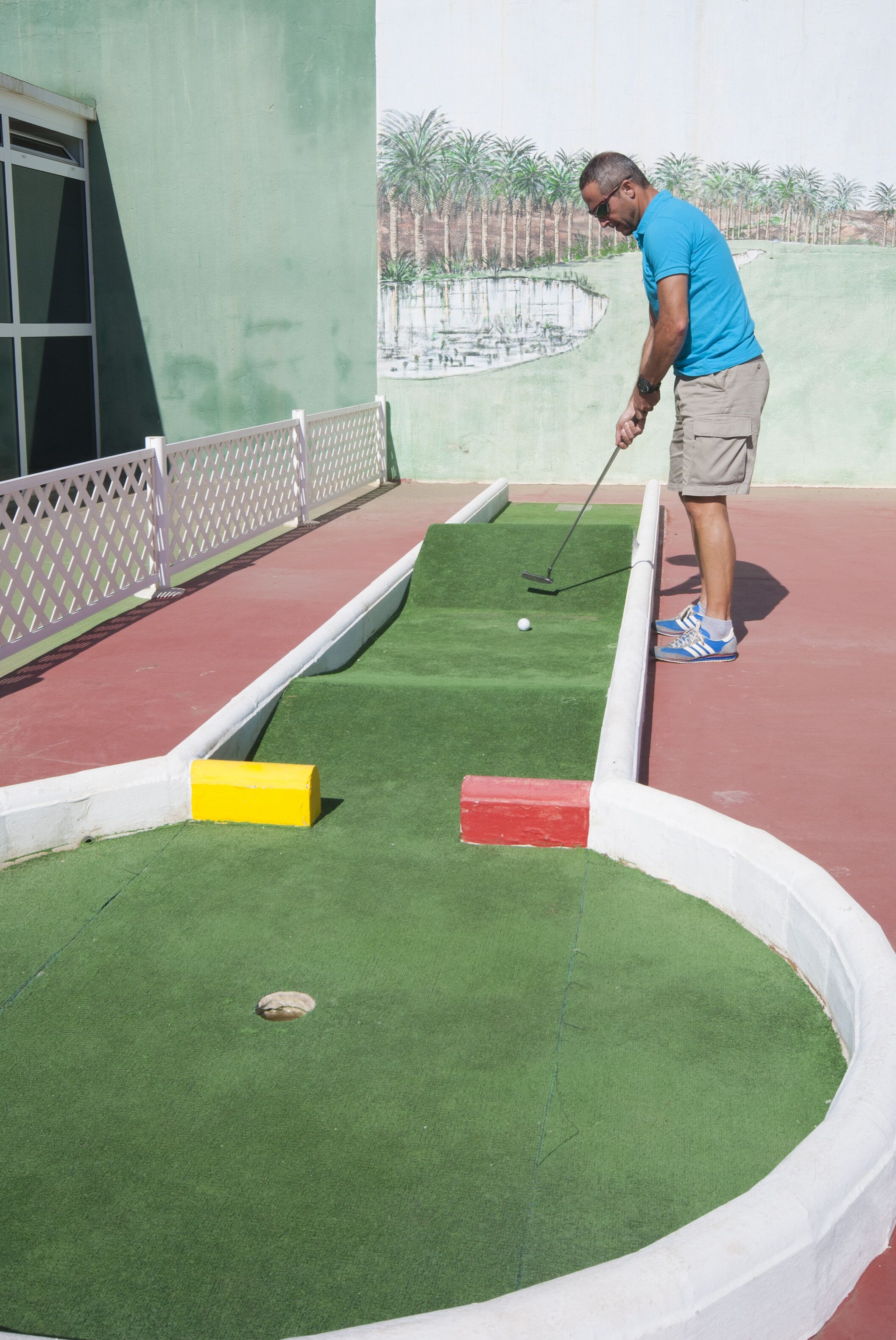 Une personne joue au croquet sur un terrain recouvert d’une pelouse artificielle verte, délimité par une ligne blanche et agrémenté d’un bloc jaune et rouge.