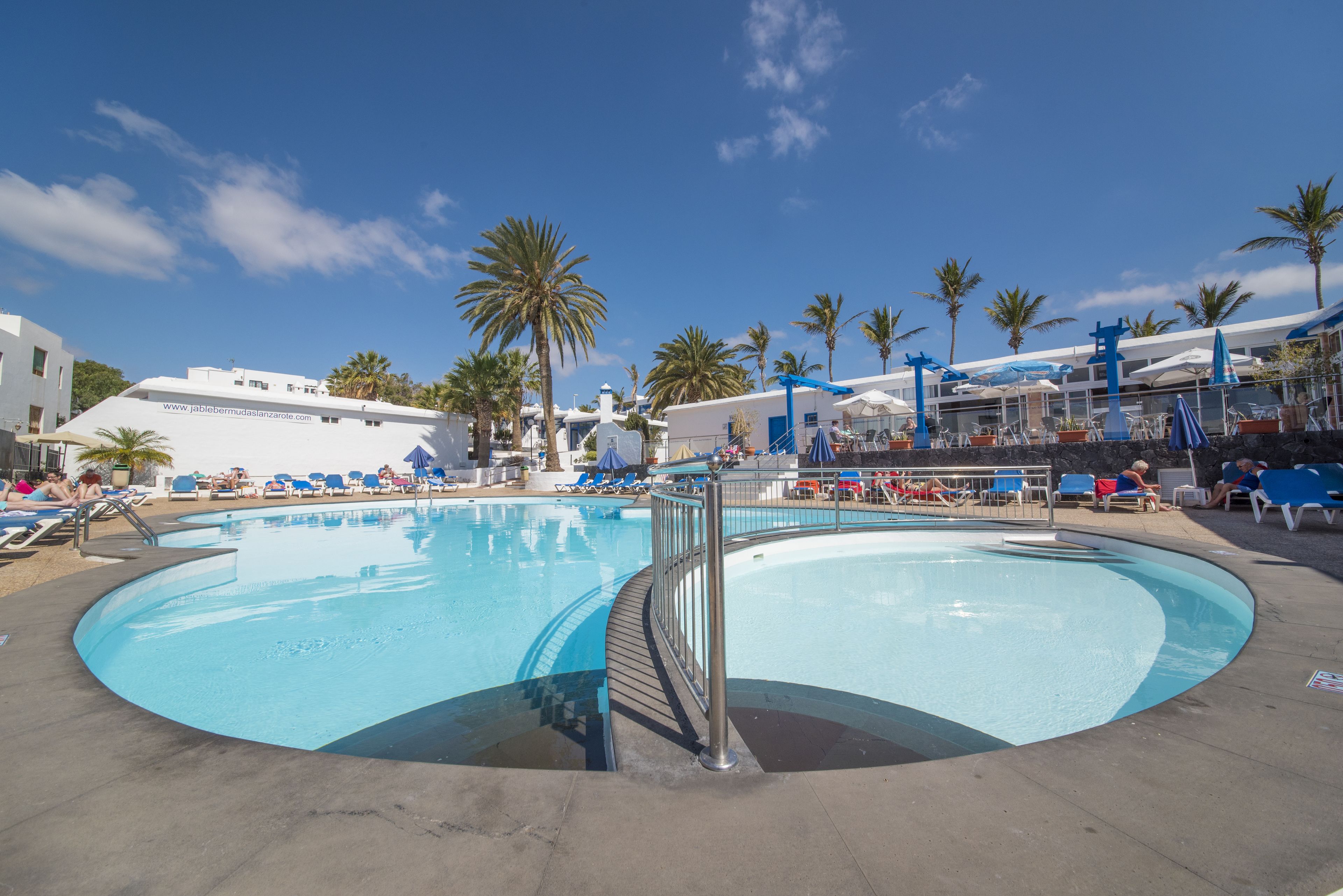 A large outdoor swimming pool with lounge chairs and umbrellas, surrounded by palm trees and a building with a white facade.