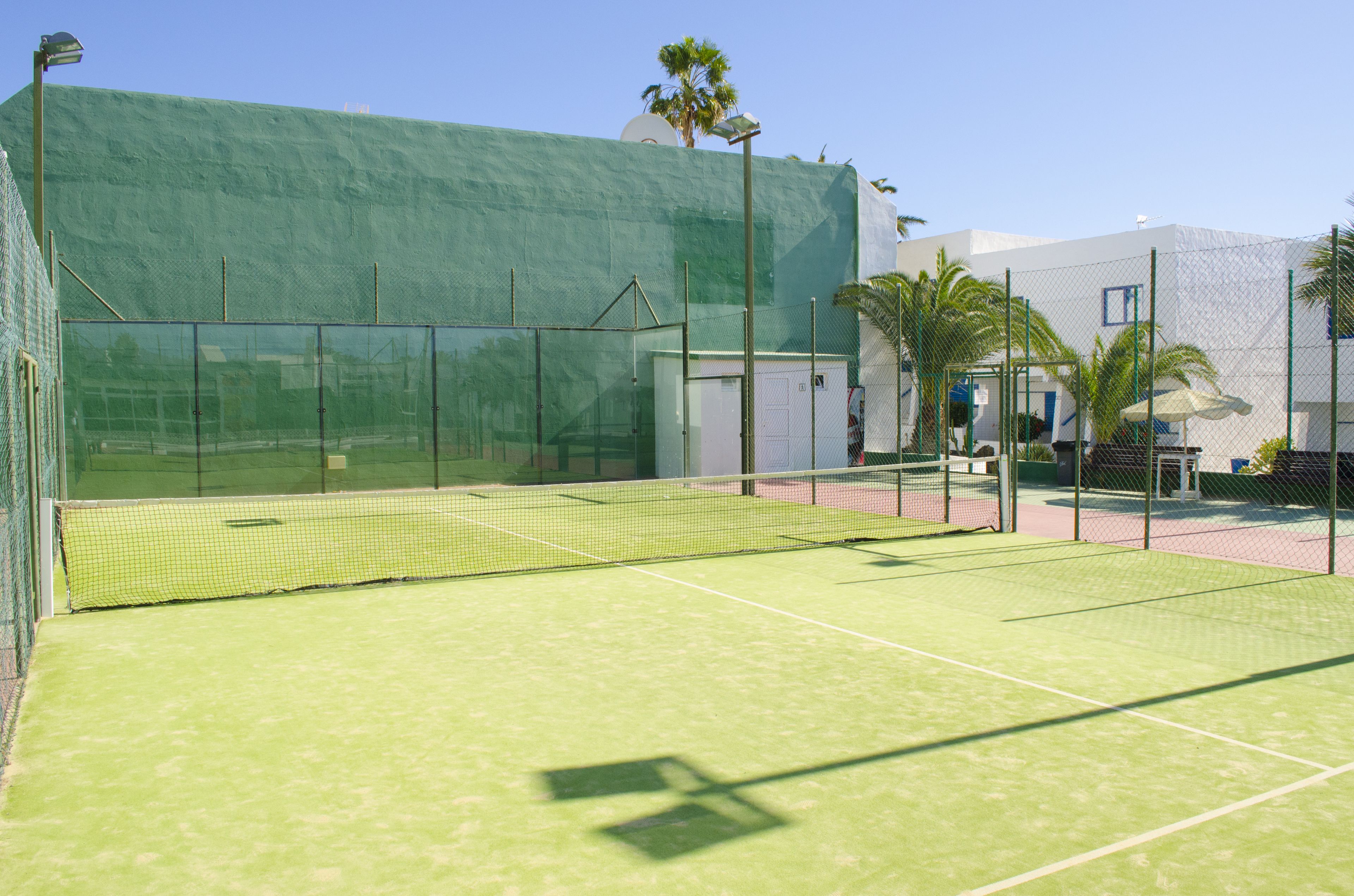 Una cancha de tenis con superficie verde, rodeada por una cerca de malla metálica y contigua a un edificio con fachada blanca y techo verde.