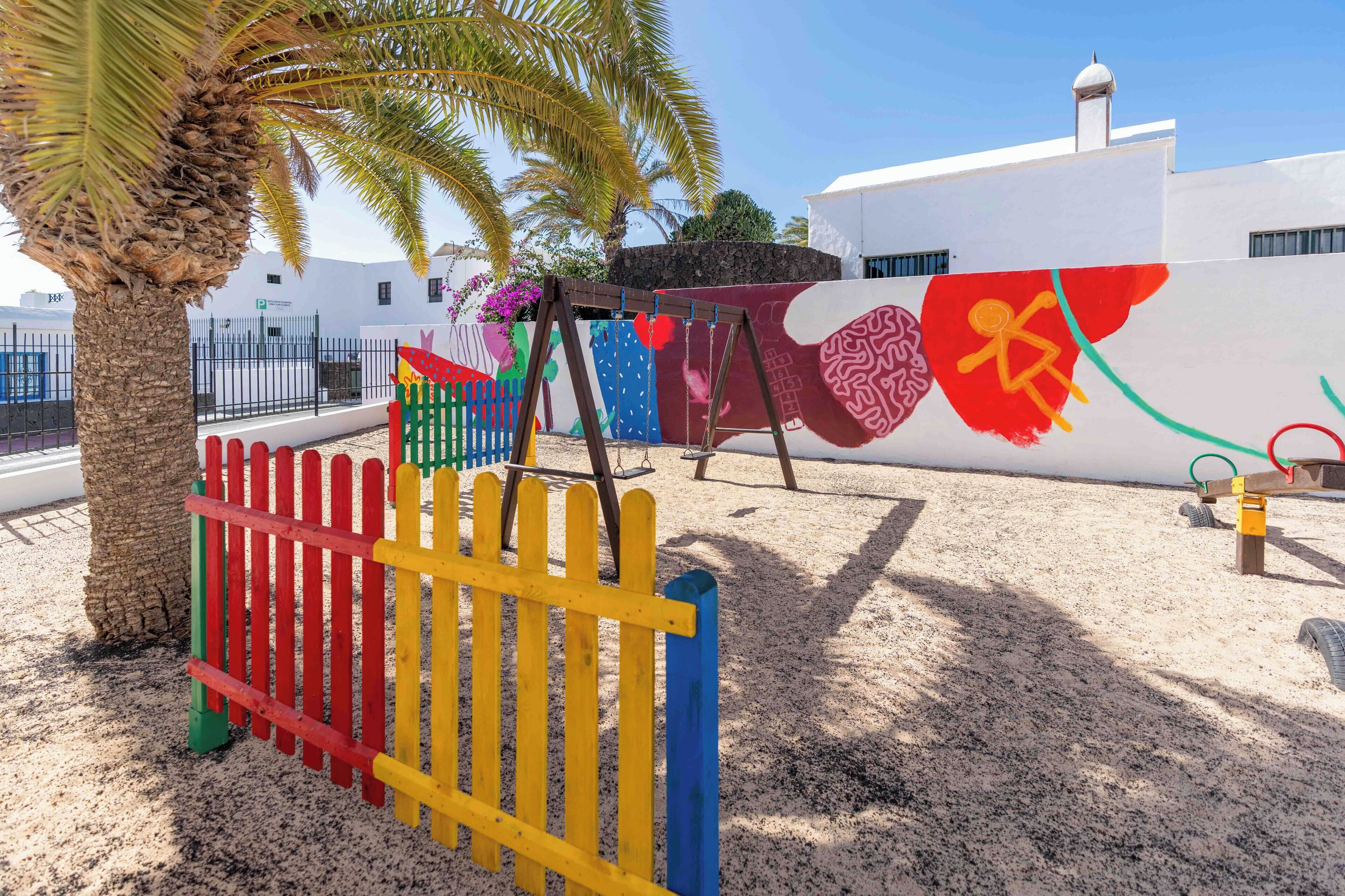 Un parque infantil con estructuras de juego coloridas, una palmera y un edificio con un mural al fondo.
