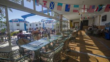 An outdoor cafe with tables and chairs, flags hanging from the ceiling, and a view of the street outside.