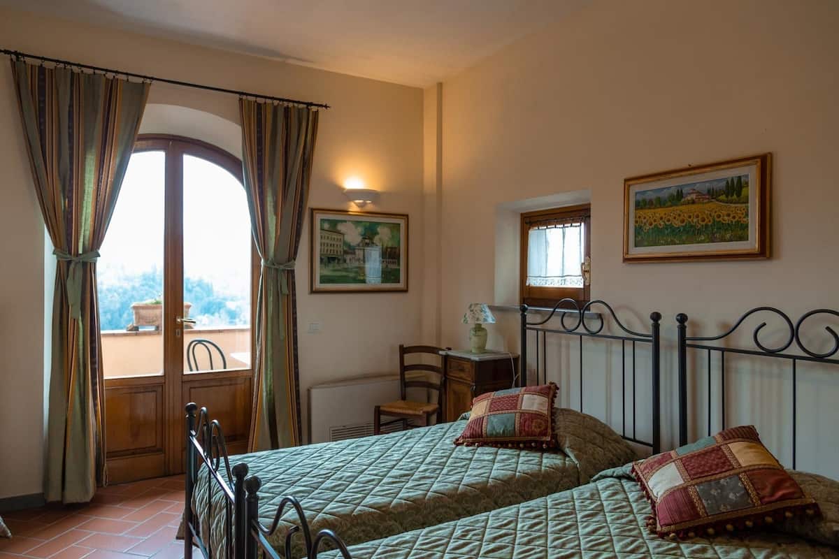 A bedroom with two beds, a window with curtains, a radiator, and two paintings on the wall.