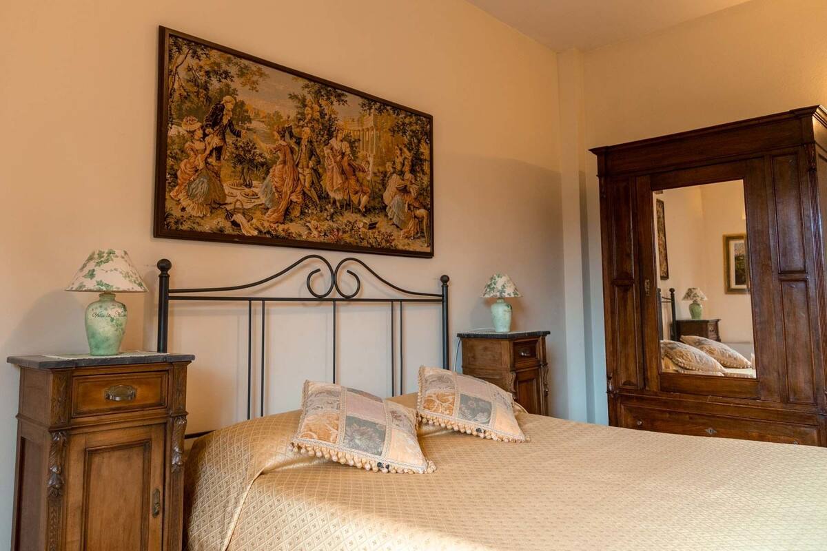 A bedroom with a classic design, featuring a large bed with ornate headboard, two bedside tables with lamps, a large mirror, and a detailed tapestry on the wall.