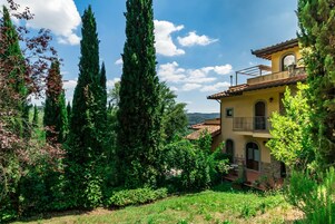 Una casa a due piani con un balcone, circondata da alti cipressi e rigogliosa vegetazione.