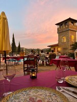 Un'area pranzo all'aperto con tavoli e sedie, una piscina e una torre in pietra sullo sfondo.