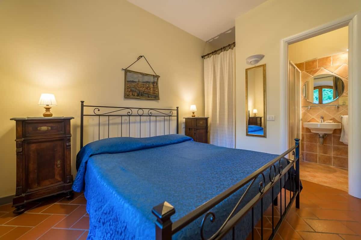 A bedroom with a bed, bedside tables, a hanging artwork, a mirror, and a bathroom with a sink and mirror.