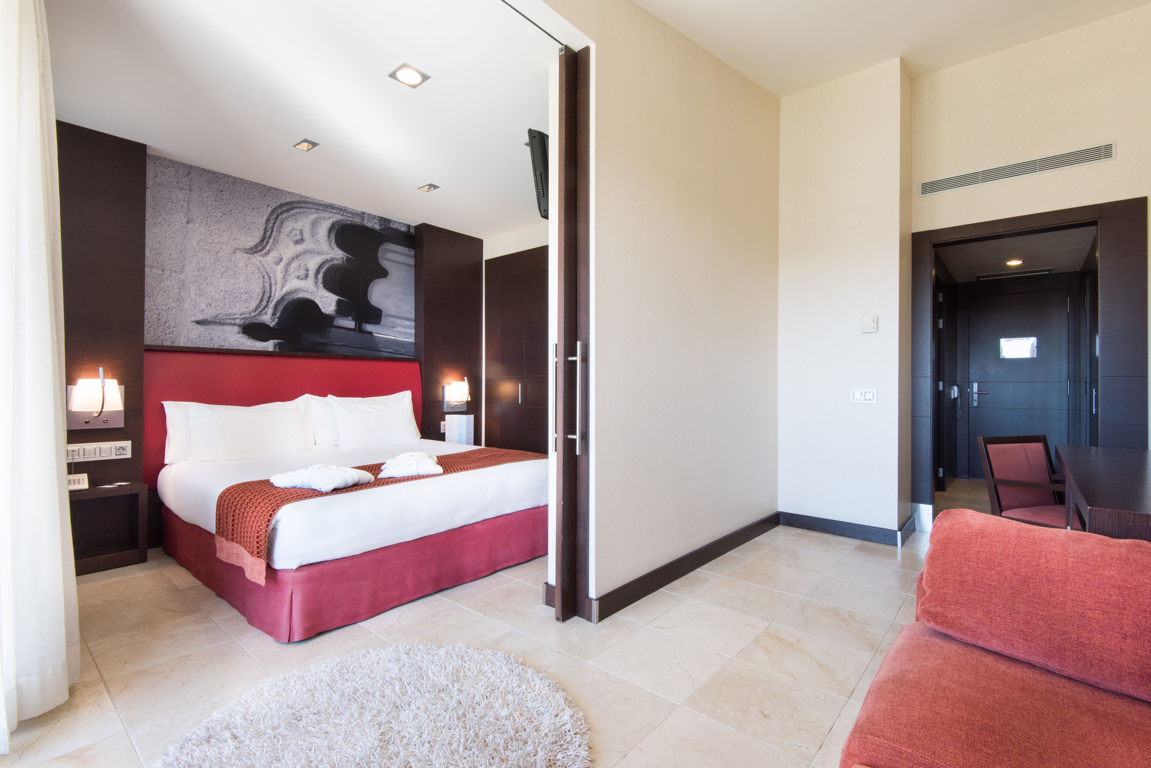 Une chambre d’hôtel moderne avec un grand lit, un mur d’accent rouge, un tableau et un coin salon séparé avec un canapé rouge.