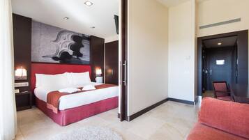 Une chambre d’hôtel moderne avec un grand lit, un mur d’accent rouge, un tableau et un coin salon séparé avec un canapé rouge.