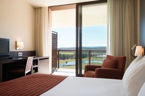 A hotel room with a large bed, a desk with a TV, a chair, and a view of a lake and mountains through a large window. - Mas Sola (Santa Coloma de Farners)