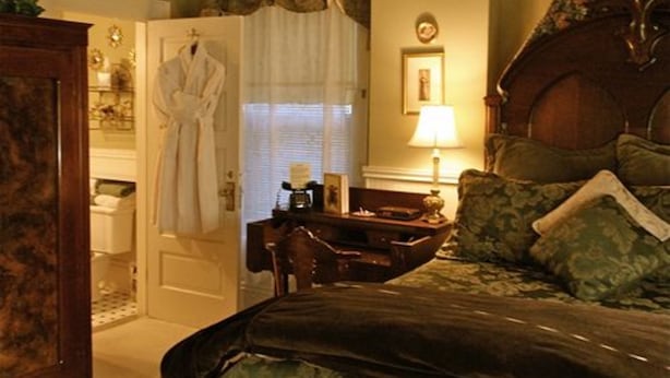 Un dormitorio con cama, escritorio y silla. Se ve una ventana con cortinas, una lámpara y un cuadro en la pared.
