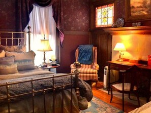 A bedroom with a bed, a chair, a desk, and a window with stained glass. - Brass Pineapple Inn (Charleston)