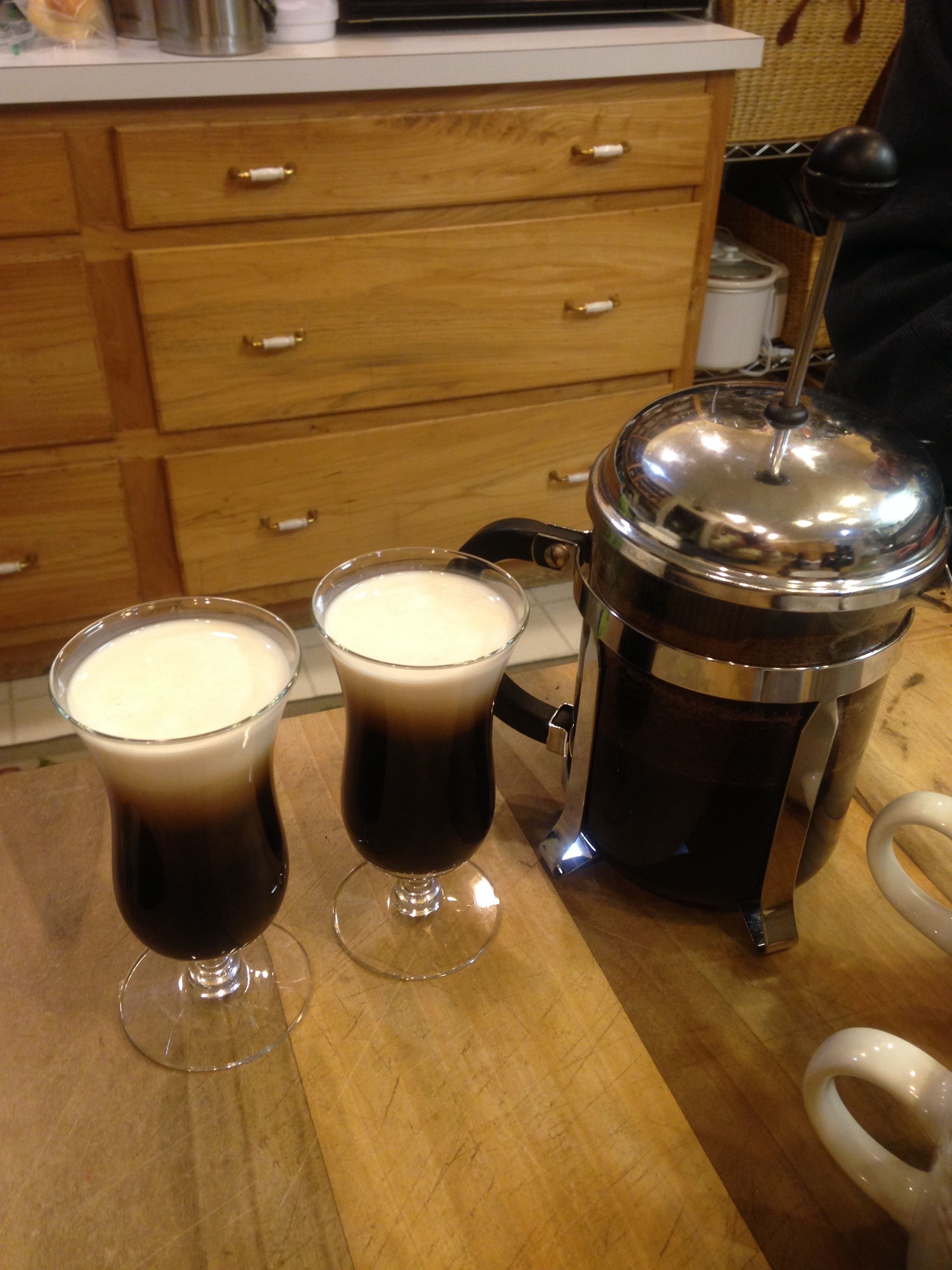Two glasses of dark liquid on a wooden surface, a metal container with a lid, and a white object with a handle.