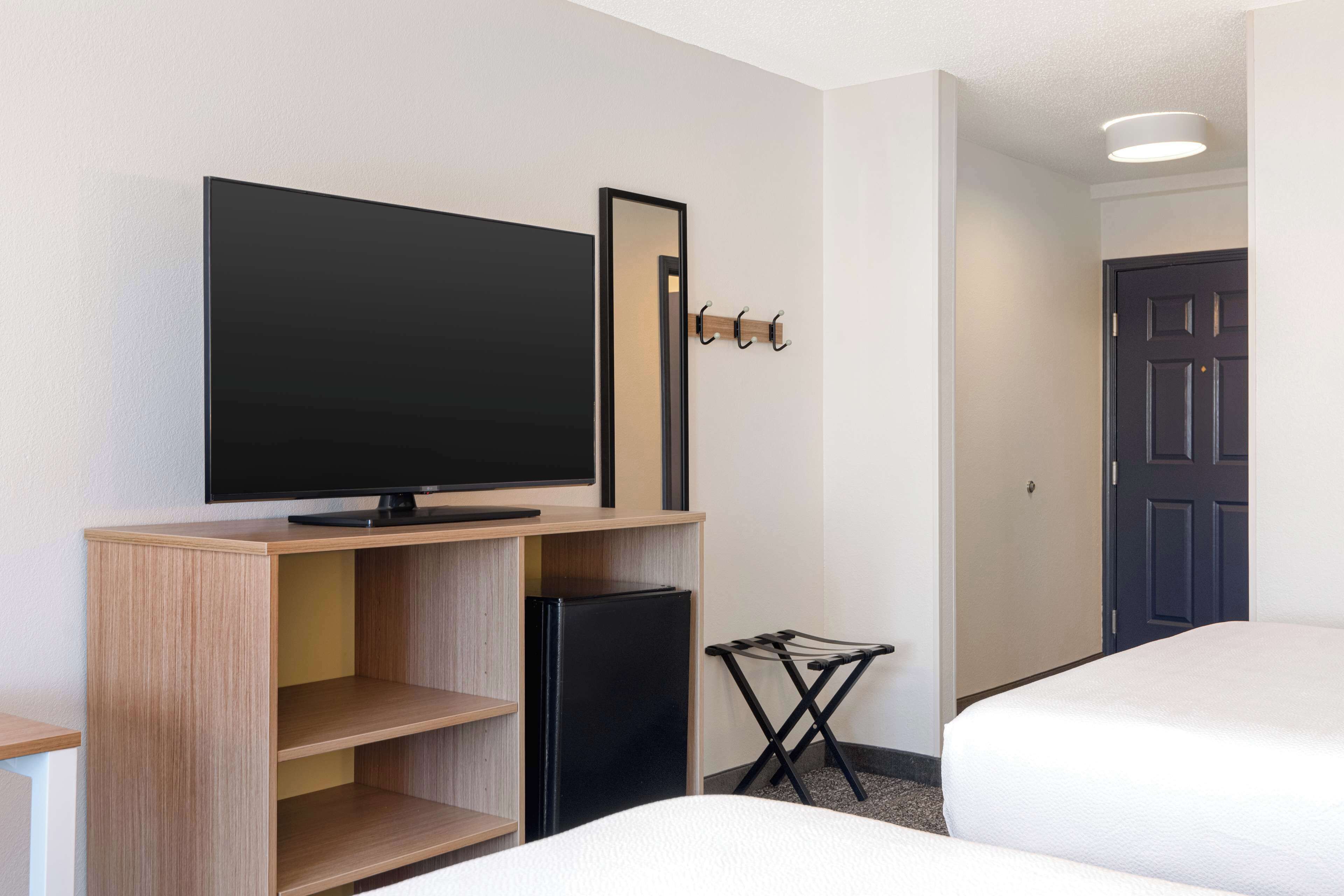 A hotel room with a flat-screen TV on a wooden stand, a small refrigerator, a coat rack, a folding chair, and two beds.