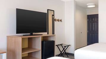 A hotel room with a flat-screen TV on a wooden stand, a small refrigerator, a coat rack, a folding chair, and two beds.
