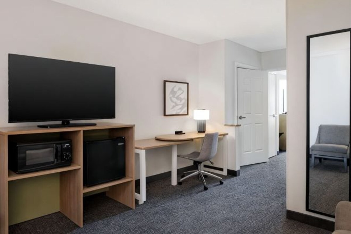 Een hotelkamer met een flatscreen-tv, een bureau met lamp, een magnetron, een stoel en een grote spiegel.