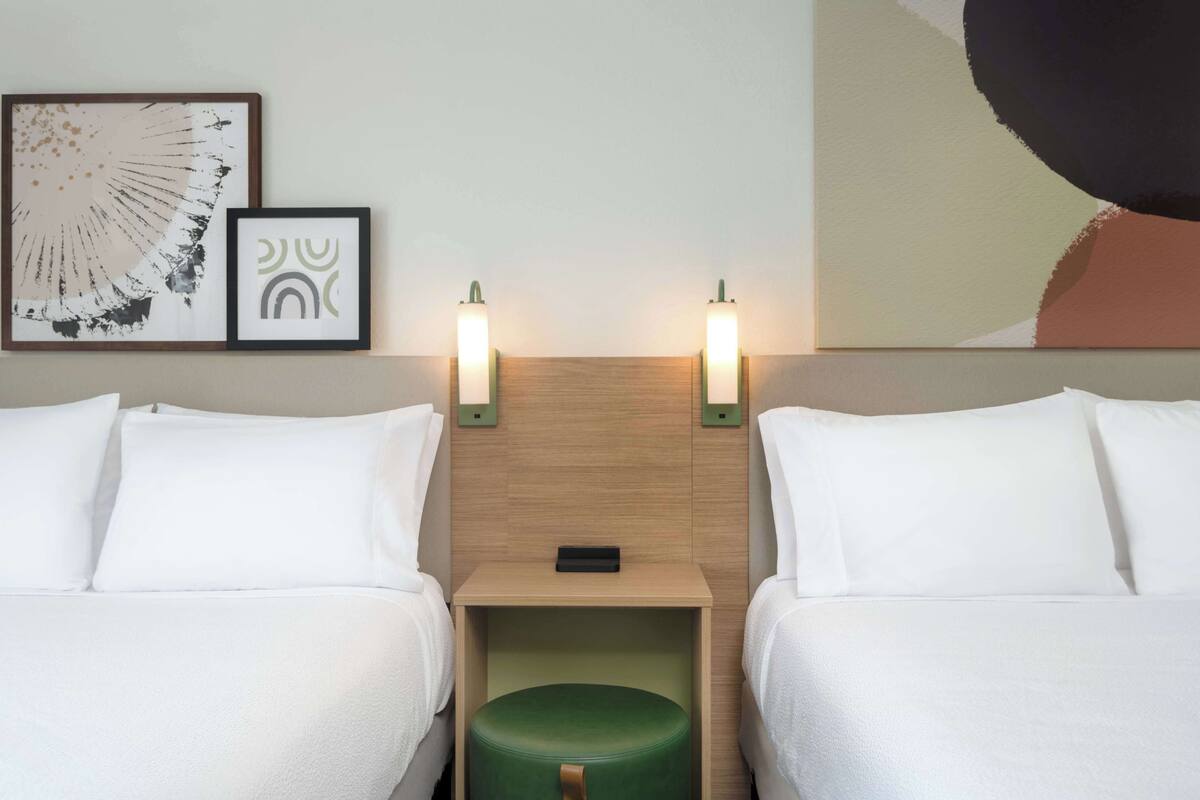 Habitación de hotel con dos camas, un reposapiés verde, cabecera de madera y dos lámparas fijadas a la pared.