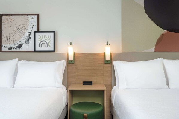 Quarto de hotel com duas camas, um banquinho verde, uma cabeceira de madeira e duas luminárias fixadas na parede.