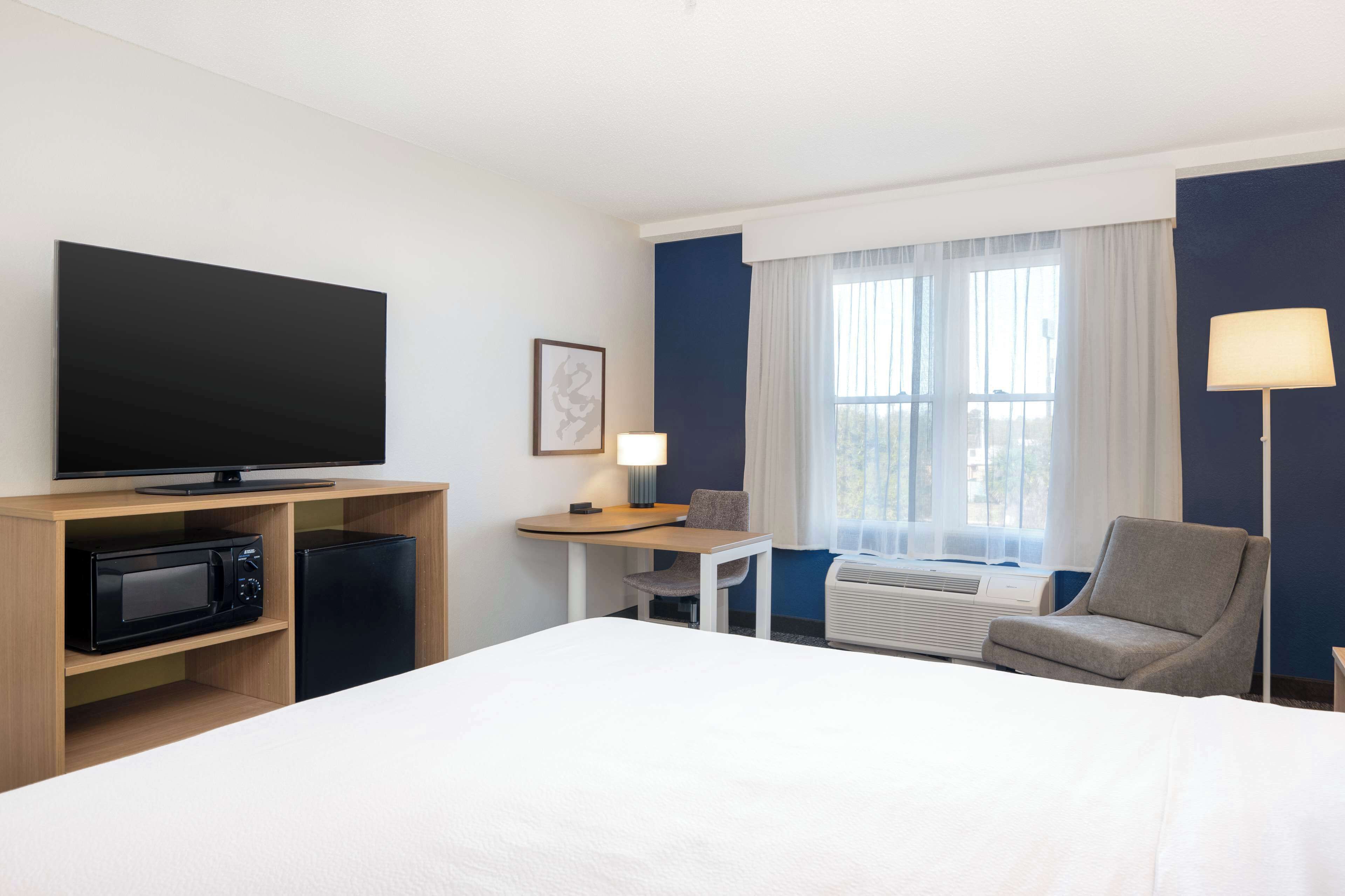 Habitación de hotel con una cama, televisión, escritorio con lámpara, silla, microondas y refrigerador.