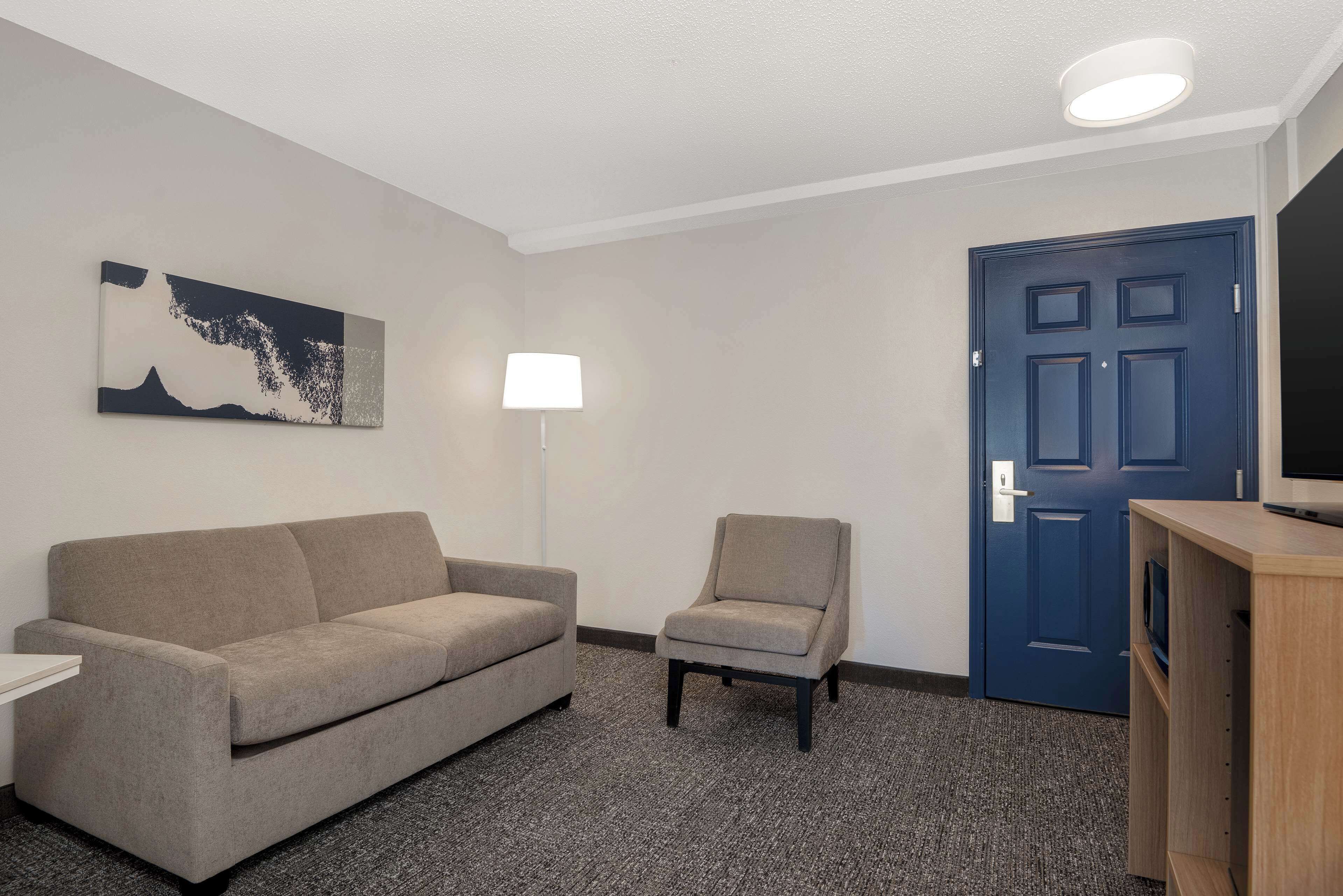 A hotel room with a grey sofa, a small armchair, a TV on a wooden stand, a blue door, and a painting on the wall.