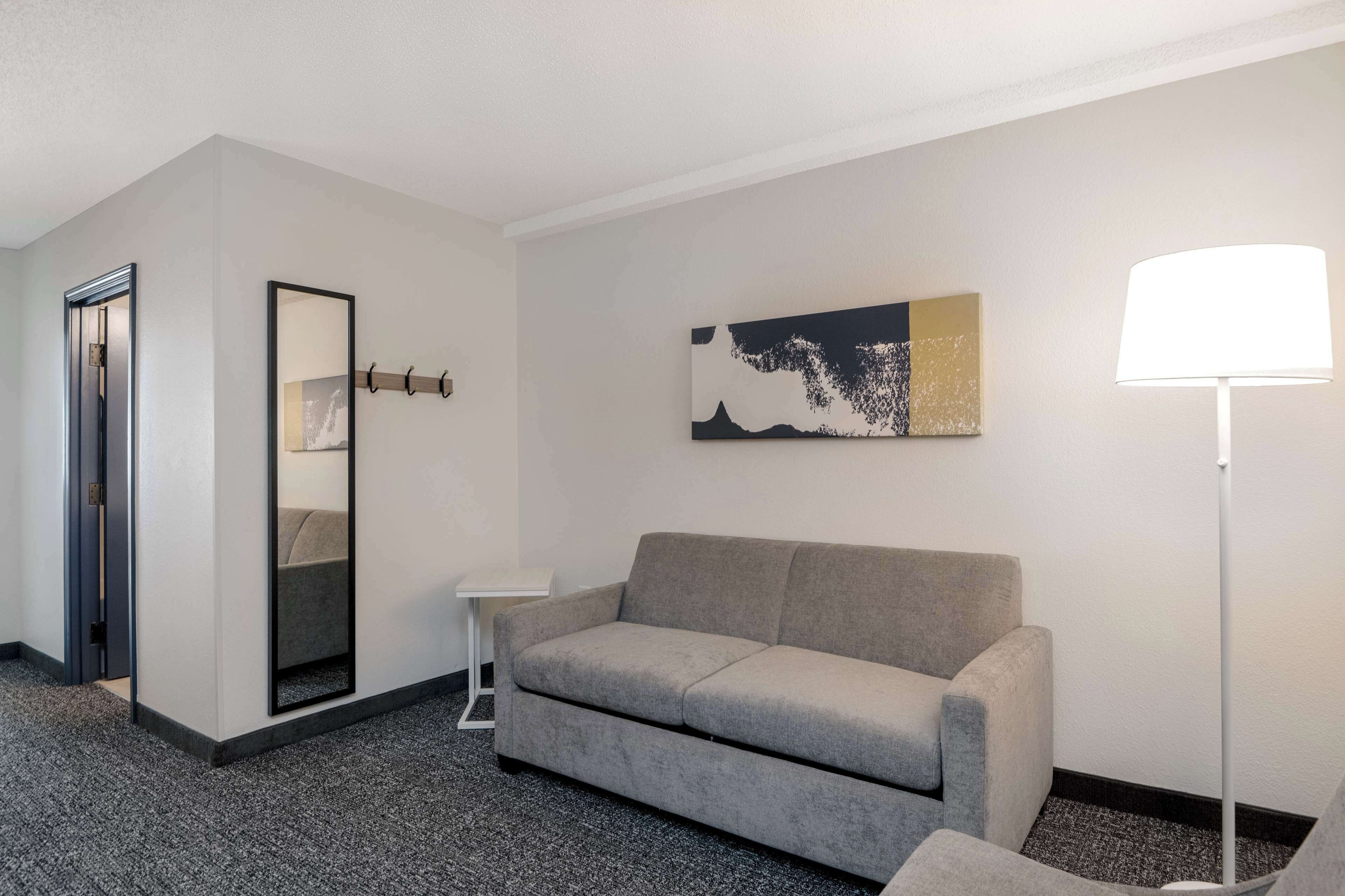 Quarto de hotel moderno com sofá cinzento, mesinha de apoio branca, luminária de chão, espelho grande e um quadro na parede.