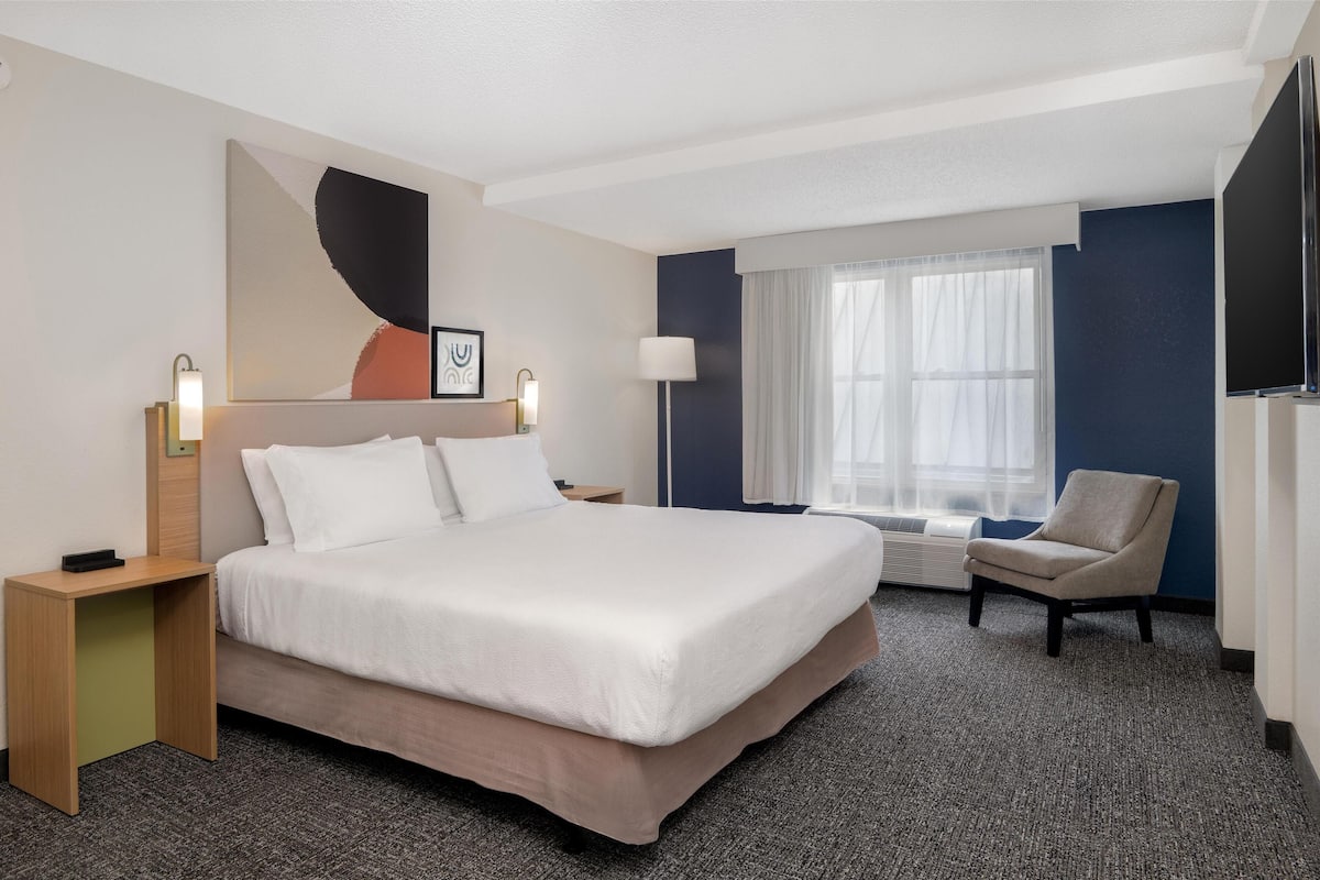 Habitación de hotel con una cama grande, un escritorio, una silla, un televisor de pantalla plana y un ventanal con cortinas.