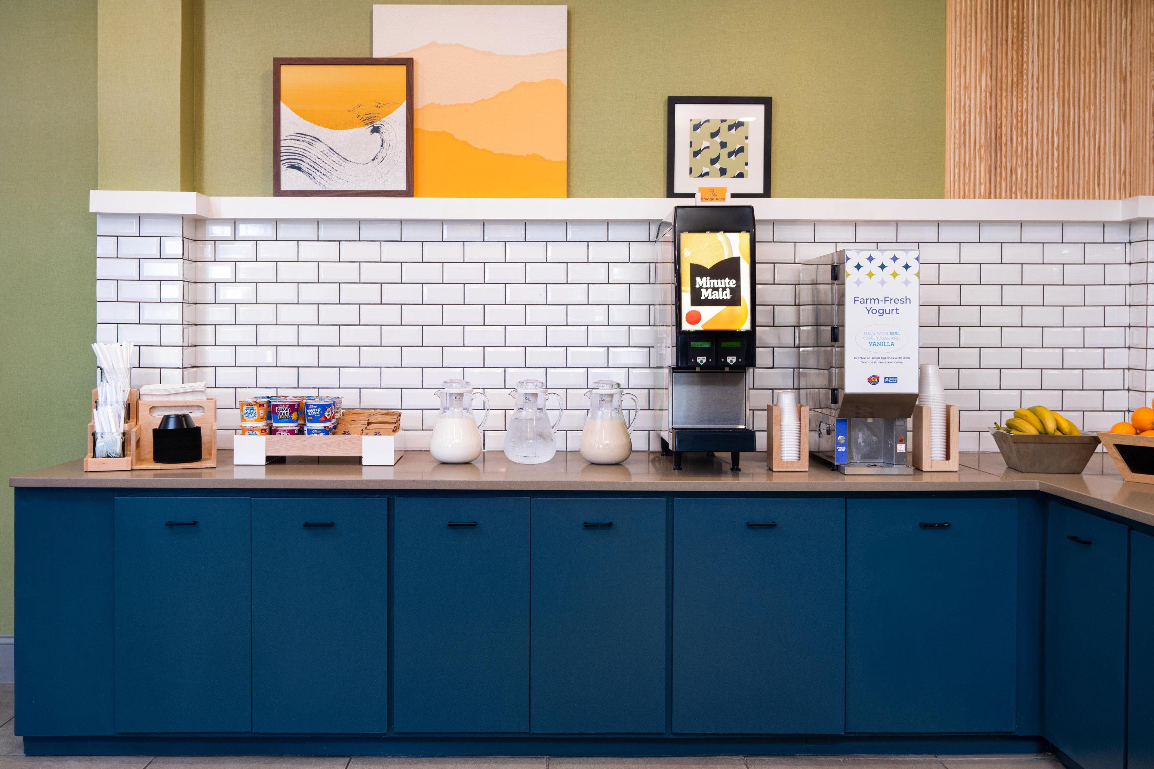 Un coin cuisine équipé d’une machine à café, d’un distributeur de lait et d’un distributeur de yaourt. On y trouve également des placards, un plan de travail et un mur décoré d’œuvres d’art.